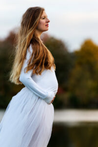 Schwangere Frau in einem weißen Kleid steht am Ufer eines Sees und genießt die ruhige Natur.