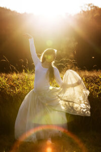 Eine schwangere Ballerina tanzt im hohen Gras, ihr weißes Kleid leuchtet im Sonnenaufgang.