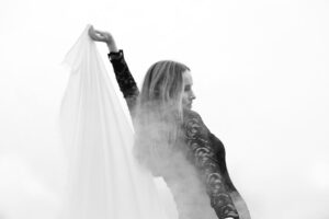 Eine Frau in einem schwarzem Kleid bewegt sich elegant im Nebel, während sie ein Tuch in der Hand hält.