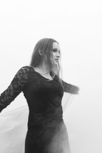 Eine Frau in einem schwarzen Kleid hält einen weißen Schal in der Hand und tanzt im Nebel.
