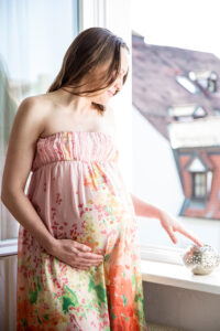Eine schwangere Frau in einem bunten Blumenkleid blickt lächelnd aus dem offenen Fenster und genießt den Augenblick der Stille.