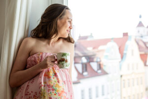 Eine schwangere Frau in einem bunten Blumenkleid blickt lächelnd aus dem Fenster und hält eine Tasse Tee in der Hand