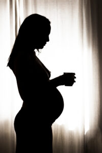 Silhouette einer schwangeren Frau mit einer Tasse in der Hand vor einem Fenster, das warmes Licht in den Raum strömen lässt.