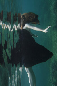 Eine Frau in einem schwarzen Kleid schwebt elegant unter der Wasseroberfläche, umgeben von blauen Wellen und Lichtreflexen.