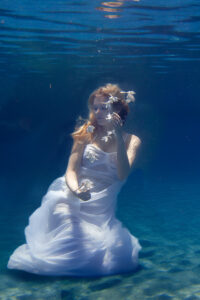 Eine Frau in einem weißen Kleid kniet elegant unter Wasser, umgeben von weißen Blüten die an die Wasseroberfläche schweben.