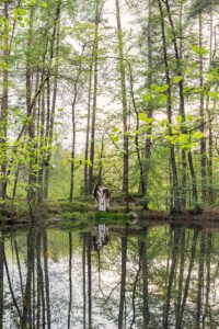 Eine Frau im weißen KLeid steht mit einem Pferd vor einem Waldsee, die Bäume spiegeln sich im Wasser.