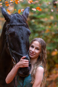 Eine Frau posiert mit einem Pferd im Wald, umgeben von Bäumen und natürlichem Licht.